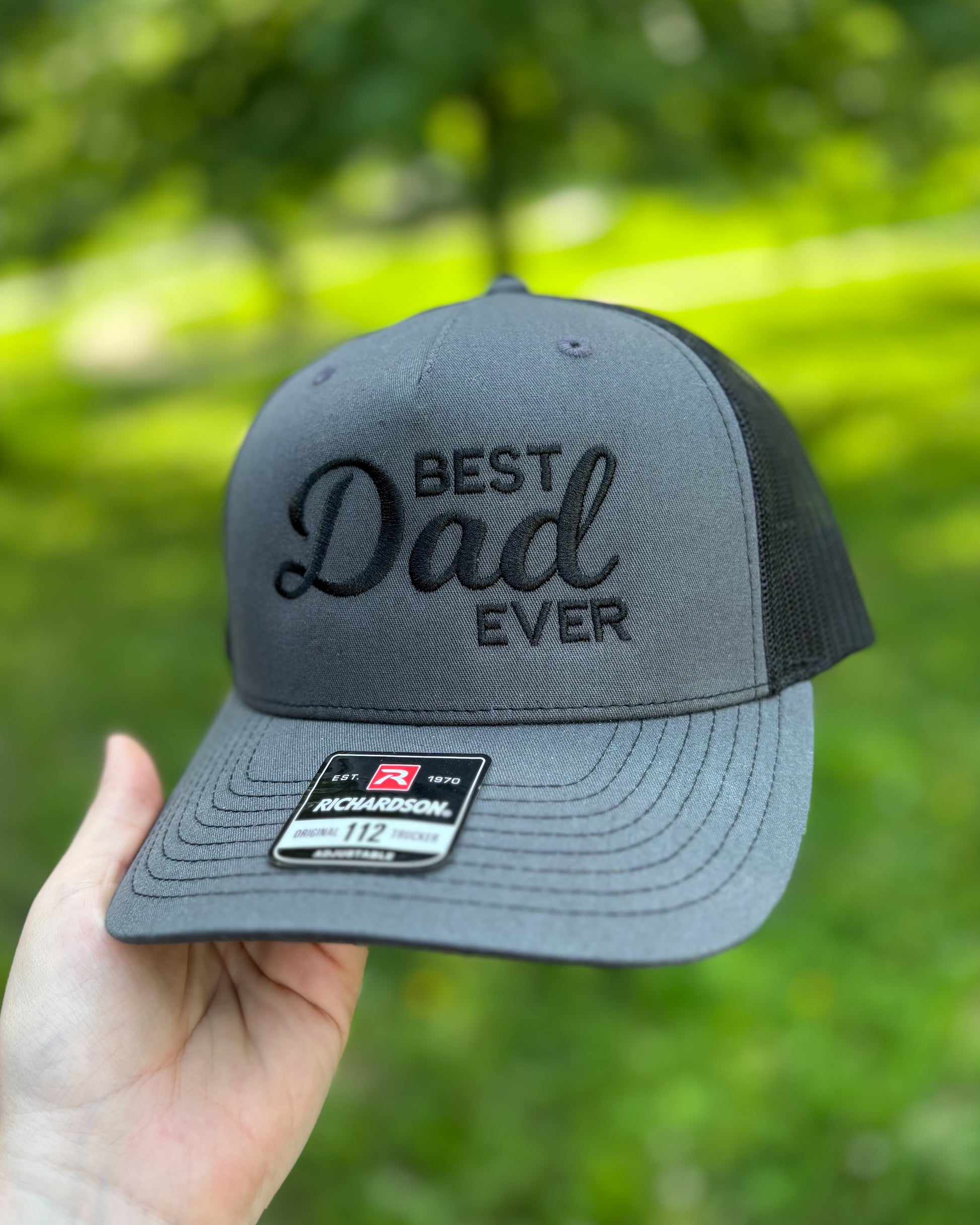 Gray Richardson 112 trucker hat with black embroidery reading “Best Dad Ever,” held in a hand outdoors against a blurred green background. The hat features a structured front panel, mesh back, curved brim, and monochromatic black-on-gray design.