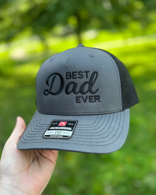 Gray Richardson 112 trucker hat with black embroidery reading “Best Dad Ever,” held in a hand outdoors against a blurred green background. The hat features a structured front panel, mesh back, curved brim, and monochromatic black-on-gray design.