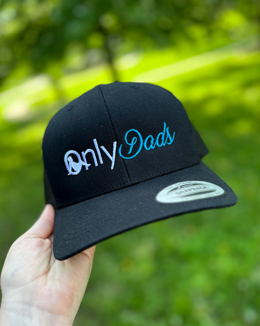 Black cap with 'Only Dads' text held in front of a blurred green outdoor background