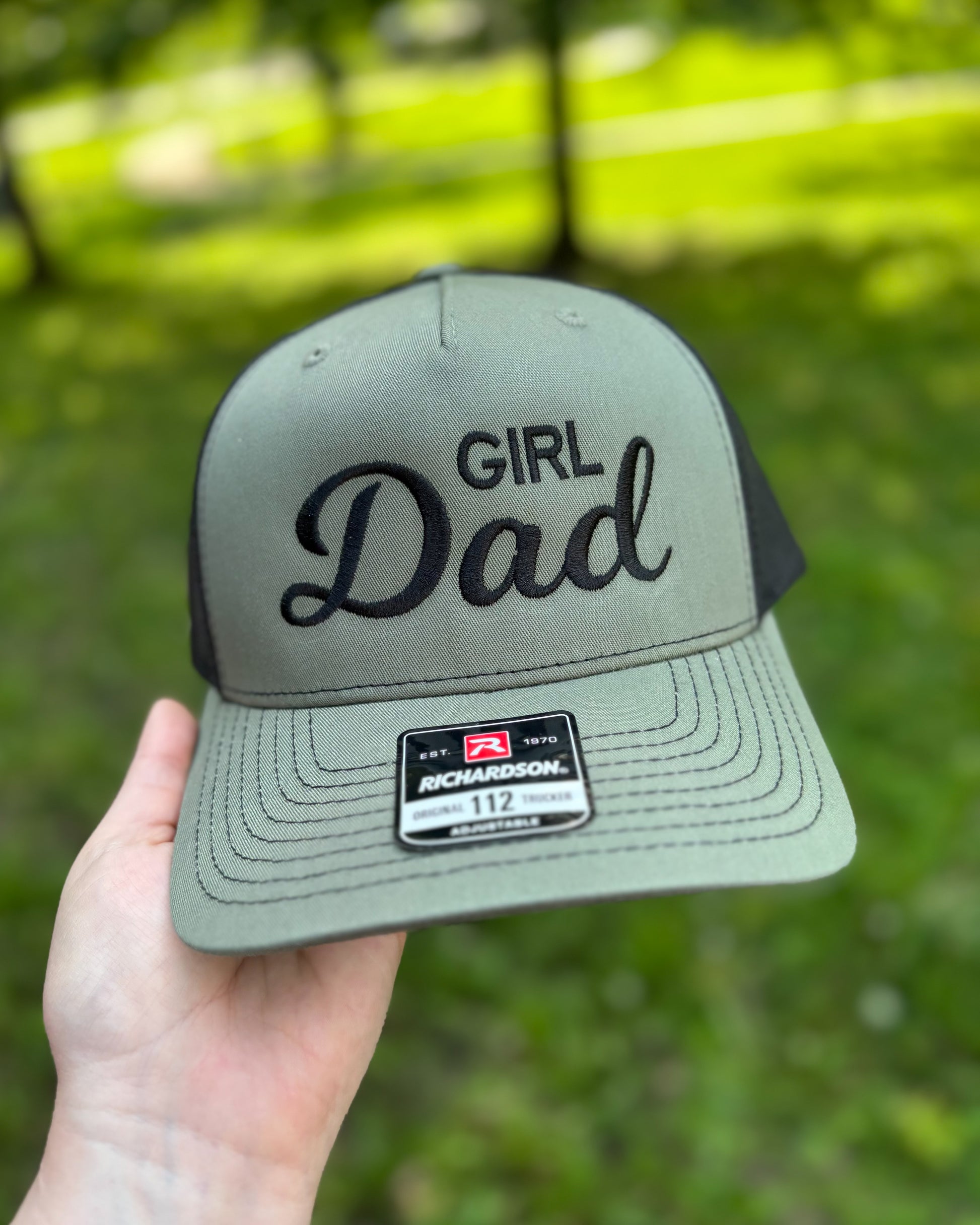Green cap with 'Girl Dad' text held by a hand against a blurred green background