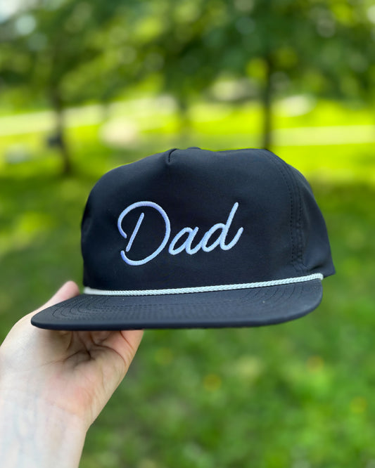 Black cap with 'Dad' embroidered on it held in front of a blurred green outdoor background