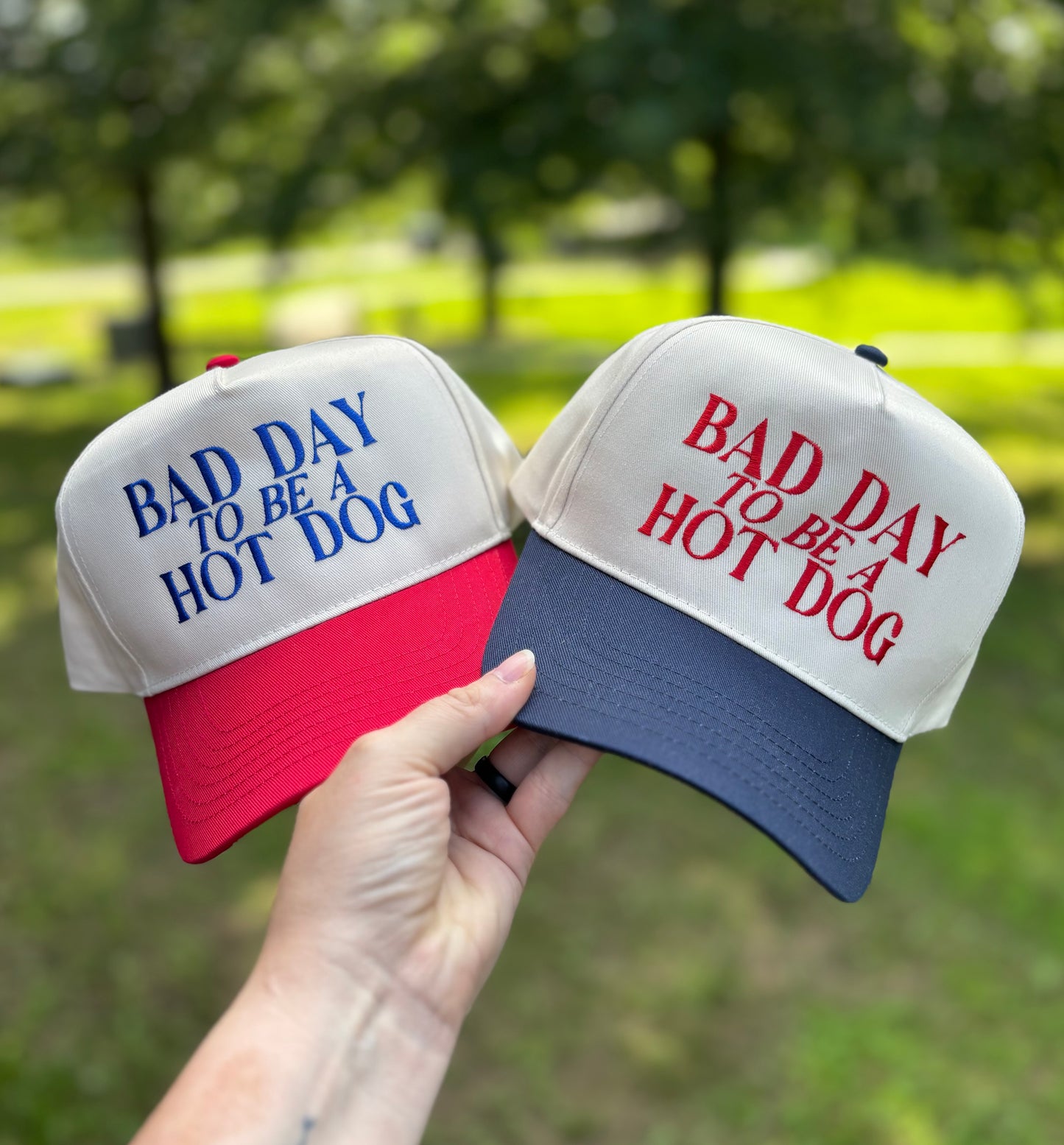 Two baseball caps with 'Bad Day to be a Hot Dog' text, one red and white, one blue and white, held against a blurred green outdoor background.