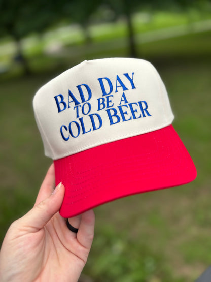 Red and white cap with 'Bad Day to be a Cold Beer' text held in a hand outdoors.