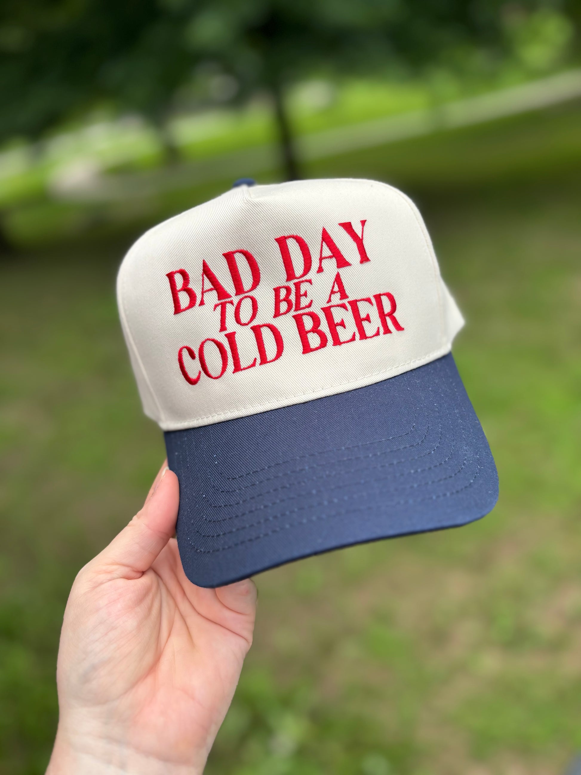 Cap with 'Bad Day to Be a Cold Beer' text held in front of a blurred green background
