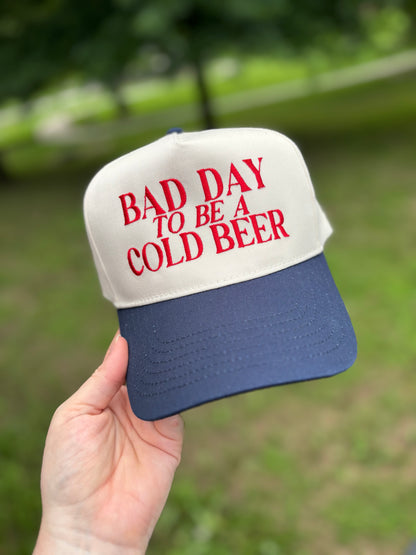 Cap with 'Bad Day to Be a Cold Beer' text held in front of a blurred green background